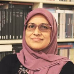 Portrait of  Sufia Azmat. She is wearing a mauve hijab with a black sweater. Books are in the background. She is wearing glasses